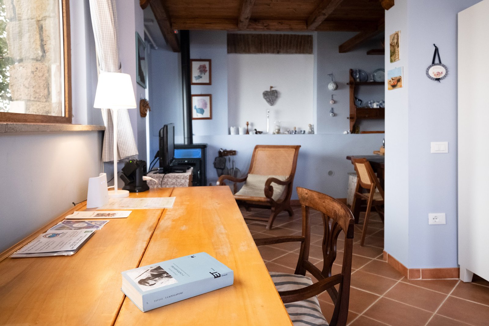 Scrivania della stanza  da letto con un libro, di fronte ad una grande vetrata e sullo sfondo la stufa a legna con poltrona e tv